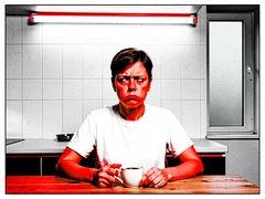 A person in a plain white t-shirt sitting at a kitchen table with a cup of coffee, staring blankly ahead with a deadpan expression. Harsh overhead kitchen strip light, flat even illumination, no mood lighting. White tiled wall behind, a single window to the right letting in flat grey daylight. Atmosphere is dry and absurdist. Raw photocopy zine aesthetic, brutalist DIY print energy with high-contrast pure white #FFFFFF and pure black #000000, disrupted by aggressive red #FF0000 accents and muted grey #F2F2F2 mid-tones. Flat hard daylight with crushed shadows and no soft falloff. Style: brutalist zine. Mood: confrontational.