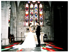 A bride and groom standing at the altar of a grand stone church, dressed in a white gown and black tuxedo, facing each other with hands joined. Tall stained-glass windows behind them cast geometric coloured light across the floor. Harsh side-lit flash from the left, deep black shadows on the right. Atmosphere is ceremonial and stark. Raw photocopy zine aesthetic, brutalist DIY print energy with high-contrast pure white #FFFFFF and pure black #000000, disrupted by aggressive red #FF0000 accents and muted grey #F2F2F2 mid-tones. Flat hard daylight with crushed shadows and no soft falloff. Style: brutalist zine. Mood: confrontational.