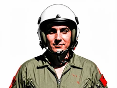 A military pilot in an olive flight suit and a white helmet with the visor raised stands facing the camera, face fully visible with a calm confident expression, oxygen mask unclipped and hanging at the chin, plain white background, flat daylight from directly above casting hard black shadows under the helmet rim and collar. Raw photocopy zine aesthetic, brutalist DIY print energy with high-contrast pure white #FFFFFF and pure black #000000, disrupted by aggressive red #FF0000 accents and muted grey #F2F2F2 mid-tones. Flat hard daylight with crushed shadows and no soft falloff. Style: brutalist zine. Mood: confrontational.