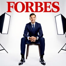 A confident businessperson in a sharp navy suit sits on the cover of a glossy financial magazine, bold red masthead reading 'FORBES' in capital serif letters at the top, clean white studio background, soft even lighting from large softboxes on both sides, polished shoes and a subtle smile conveying success. Style: business magazine cover photography. Mood: ambitious.