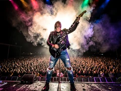 A rock musician in a leather jacket and ripped jeans performs on a massive concert stage, electric guitar raised high, crowd of thousands stretching into darkness below, dramatic backlit smoke and multicolored stage spotlights creating a halo effect, sweat and energy visible in every detail. Style: live concert photography. Mood: electrifying.