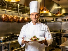 A professional chef in a crisp white double-breasted chef's coat and tall toque stands in a gleaming restaurant kitchen, holding a beautifully plated gourmet dish at chest height, stainless steel surfaces and hanging copper pots visible behind, warm overhead pendant lighting casting a golden glow on the food. Style: culinary magazine photography. Mood: confident and refined.